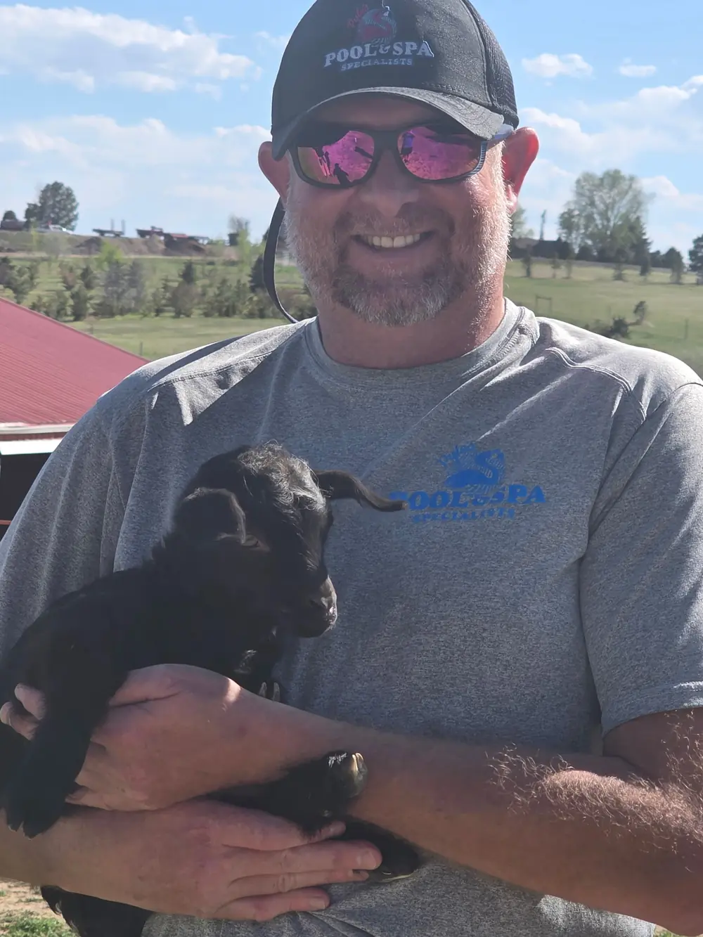 Joel, owner of Pyle's Pool & Spa Specialists, in branded gear holding a baby goat at a Colorado ranch